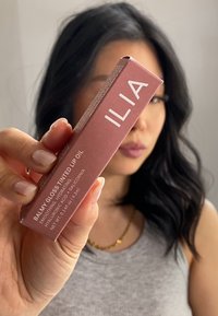 Imballaggio dell'ILIA Balmy Gloss Tinted Lip Oil in rosa opaco con testo bianco, caratterizzato da un design geometrico e informazioni sul prodotto su entrambi i lati.