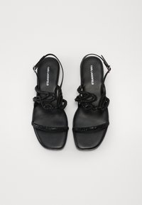 Musta slingback-sandaalit, joissa on teksturoidut hihnat, joissa on kohotettu kuvio, ja kiiltävä nauha edessä. Litistetty pohja, minimalistinen tyyli.