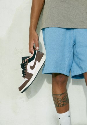 Person trägt blaue Shorts und weiße Socken, hält einen schwarzen und braunen Nike-Sneaker neben einer weißen Wand und zeigt ein Bein-Tattoo.