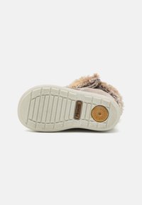 Botte en daim marron avec une semelle en caoutchouc blanc texturé, ornée d'une bordure en fourrure douce et d'un logo estampé pour une meilleure adhérence et un bon maintien.