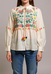 Blusa color crema con bordado floral en rojo, verde y azul. Presenta borlas y pompones coloridos, con un cierre de lazo en el cuello.