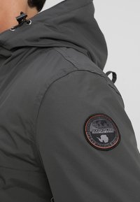 Gros plan d'une personne portant une veste gris foncé avec un écusson logo Napapijri circulaire sur le haut du bras et une capuche avec cordon ajustable.