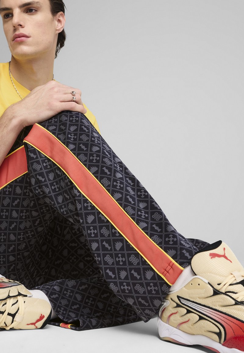 Un pantalon de jogging noir à motifs présente des rayures latérales orange et jaune. Le tissu est léger et lisse. Des chaussures jaunes avec des accents rouges sont visibles.