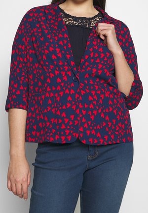 Frau trägt einen marineblauen Blazer mit roten Herzmustern über einem schwarzen Spitzen-Top und blauen Jeans, hält mit einer Hand das Revers des Blazers.
