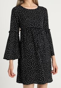 Vestido negro con mangas acampanadas, que presenta una cintura fruncida y un patrón abstracto blanco a lo largo de la tela. Textura suave.