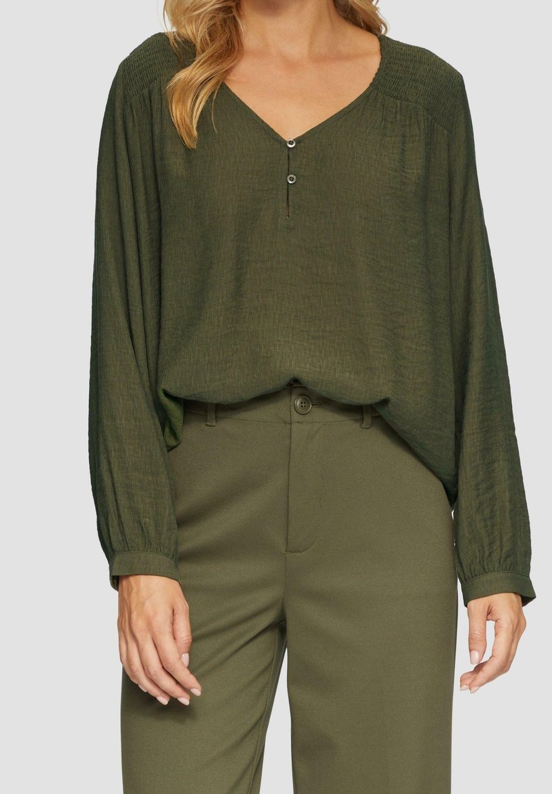 Femme portant un chemisier vert olive à manches longues avec un col en V boutonné et un pantalon taille haute assorti vert olive.