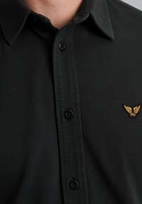 Camisa polo negra con tejido texturizado, cuello suave y botones negros. Logo de ala dorada bordado en el lado izquierdo del pecho.