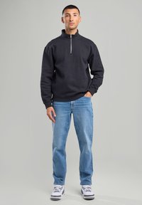 Black zip-up sweatshirt with high collar, casual fit; worn with blue denim jeans and white sneakers featuring black accents.