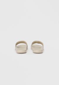 Beige slide sandals with a smooth, glossy finish. Two curved straps form the open upper, resting on a flat sole with a slight heel.