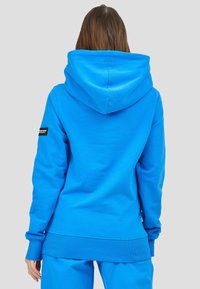 Felpa con cappuccio blu con un cappuccio grande e regolabile. Tessuto liscio, polsini e orlo a costine, con una toppa con logo sulla manica sinistra.