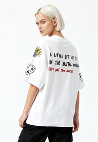 Balti per didelis marškinėliai su spalvingais siuvinėjimais saulės ir veidų ant rankovių, o nugarėlėje juodu ir raudonu tekstu.