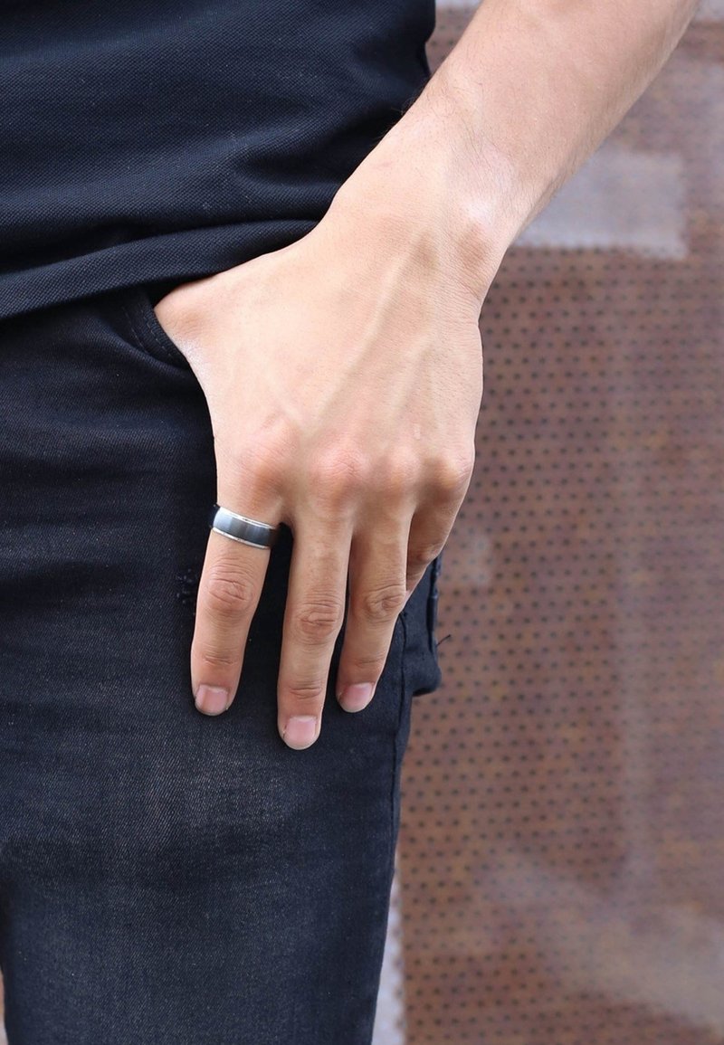 Bague en acier inoxydable portée à l'index, design plat, finition mate, sur un fond de denim texturé et de tissu.