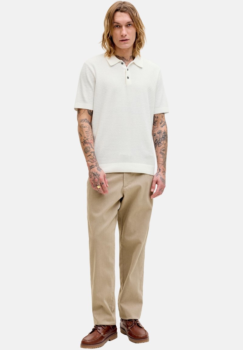 Homme tatoué portant un polo blanc à manches courtes, un pantalon beige et des chaussures en cuir marron, debout devant un fond clair uni.