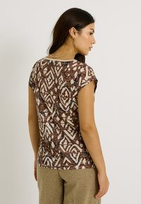 Top marrón con patrón y mangas cortas, que presenta un diseño geométrico en crema y azul, confeccionado en tejido ligero, con un escote redondeado.