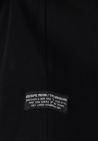 Tela negra con una etiqueta rectangular cosida que dice "ESCAPA DE / LO DESCONOCIDO", promocionando estilo y tendencias, establecida en 1994 por Key Largo.