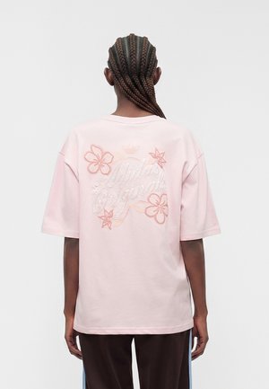 Persona con cabello trenzado lleva una camiseta rosa claro de Adidas Originals con bordado floral en la espalda, combinada con pantalones oscuros.