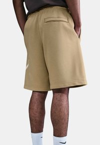 Pantalones cortos de algodón beige con cinturilla elástica, bolsillo trasero y una textura suave. Presenta un logo sutil en el lateral y un corte hasta la rodilla.