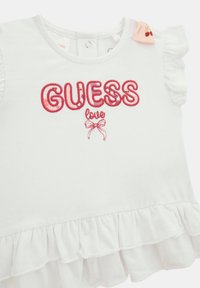 Hvid T-shirt med bølgede ærmer, prydet med rødt broderet "GUESS love" tekst og et sløjfegrafik. Blødt bomuldsstof med en flared kant.