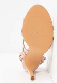 Vue de dessous d'une sandale à talons hauts beige clair avec des fines brides et un talon pointu, reposant contre une surface blanche.