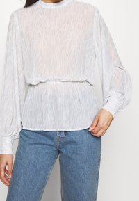 Femme portant un chemisier blanc à manches longues transparentes avec des poignets froncés et un jean en denim bleu, se tenant devant un fond uni.