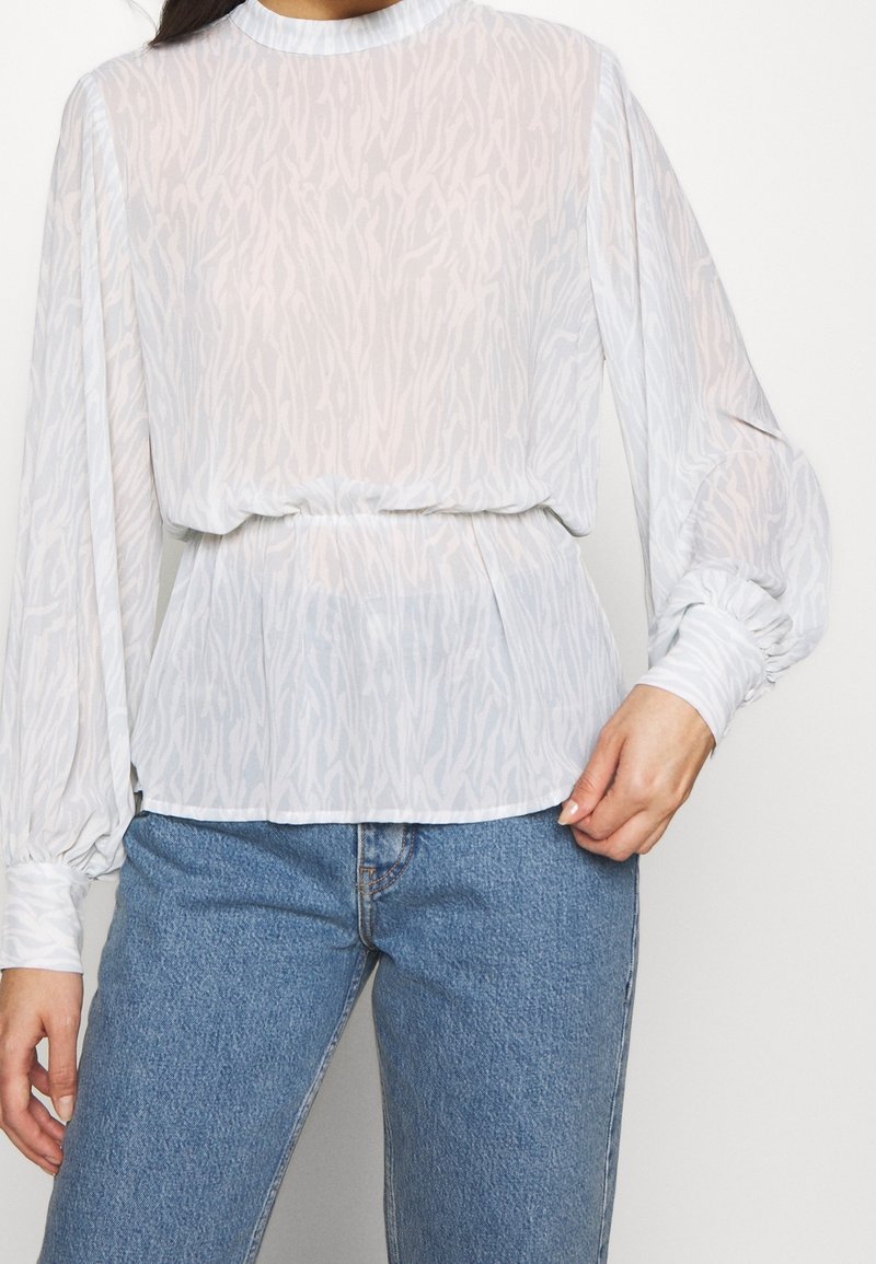 Femme portant un chemisier blanc à manches longues transparentes avec des poignets froncés et un jean en denim bleu, se tenant devant un fond uni.