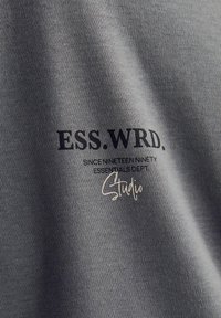 Grå bomuldsstof med trykt tekst: "ESS. WRD." i mørkeblå og "Studio" i en lysere tone, med en let ribbet tekstur.