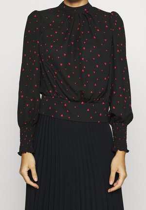 Zwarte blouse met lange mouwen en een rood hartjespatroon, gerimpelde manchetten, hoge halslijn, gecombineerd met een zwarte geplooide rok.