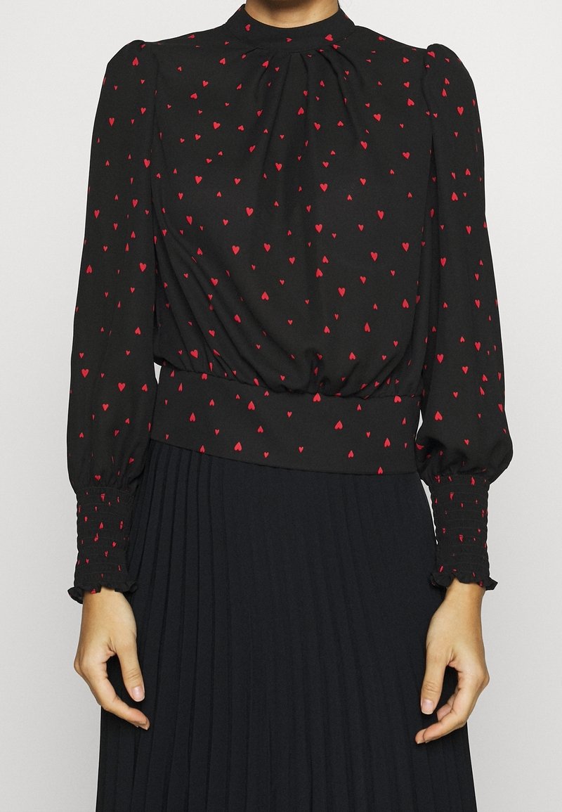 Blouse noire à manches longues avec motif de cœurs rouges, poignets froncés, col haut, associée à une jupe plissée noire.