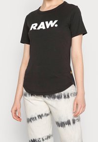 Černé bavlněné tričko s krátkým rukávem, na přední straně s velkým bílým logem "RAW." Zakulacený spodní lem a minimální akcenty.