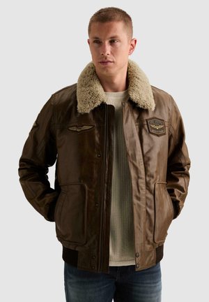 Man draagt een bruine leren jas met een grote beige shearling kraag en patches, staand met handen in de zakken tegen een effen achtergrond.