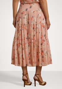 Blomstrete maxiskjørt i myk ferskenfarget stoff, med et lagdelt design med hvite og oransje blomster, og en luftig silhuett. Kombinert med høye sandaler.