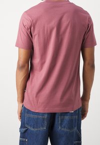 Persoon in een effen stofroze T-shirt met korte mouwen en blauwe denim cargo shorts, gezien van achteren tegen een witte achtergrond.