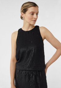 Czarny top bez rękawów z pionowymi paskami, wykonany z błyszczącego materiału. Charakteryzuje się luźnym krojem i subtelnymi cekinowymi akcentami wzdłuż pasków.