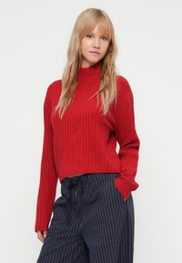 Rød ribbet genser med høy hals, med lange ermer og cropped design. Kombinert med marineblå stripete bukser.