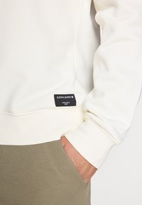 Hanorac alb cu manșete ribbate, având un eticheta de brand neagră pe partea inferioară. Asortat cu pantaloni verzi măslinii. Textură de stofă moale.