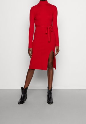 Rotes, figurbetontes Kleid mit Rollkragen und langen Ärmeln, ausgestattet mit einem seitlichen Schlitz und einem passenden Taillengürtel. Schwarze Knöchelstiefel runden den Look ab.