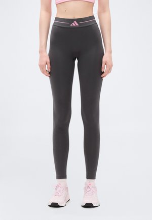 Γυναίκα που φοράει ψηλόμεσα σκούρα γκρι κολάν με ροζ λογότυπο Adidas και ροζ αθλητικά παπούτσια, όρθια μπροστά σε ανοιχτόφωτο φόντο.