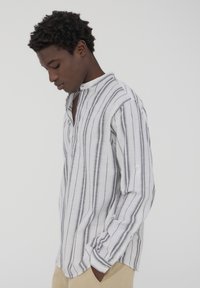 Camicia bianca a maniche lunghe con strisce verticali grigie, colletto con bottoni e vestibilità rilassata. Indossata con pantaloni beige chiaro.