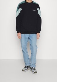 Sudadera negra con paneles en los hombros en tonos turquesa y azul claro, combinada con jeans azul claro y zapatillas multicolores.