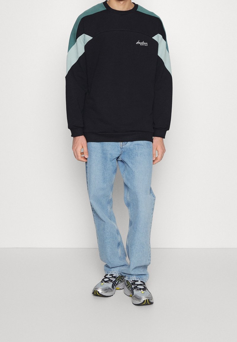 Sudadera negra con paneles en los hombros en tonos turquesa y azul claro, combinada con jeans azul claro y zapatillas multicolores.