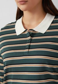 Eine Frau tr�ägt ein grün-braun-schwarz-weiß gestreiftes Poloshirt mit einem weißen Kragen und drei nah am Hals befestigten Knöpfen.