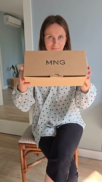 Scatola di cartone con logo "MNG" tenuta in mano. Colore marrone chiaro, forma rettangolare, lievi segni di usura sui bordi. Blusa a pois sullo sfondo.