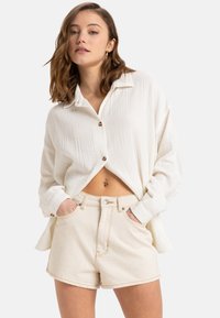Weiße strukturierte Bluse mit Knöpfen, kombiniert mit hellbeigen, hochgeschnittenen Shorts, die Fronttaschen und sichtbare Nähten aufweisen.
