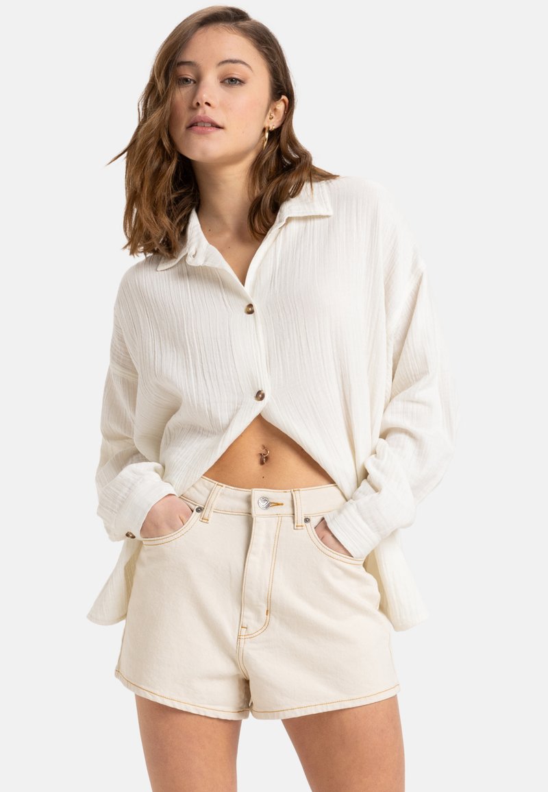 Weiße strukturierte Bluse mit Knöpfen, kombiniert mit hellbeigen, hochgeschnittenen Shorts, die Fronttaschen und sichtbare Nähten aufweisen.