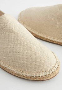 Beige canvas slip-on shoes with a rounded toe, featuring a textured upper and a natural jute trim around the sole.