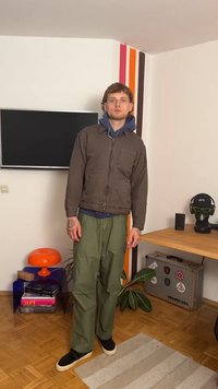 Rjava zip-up jakna, modra kapuca spodaj, zelene cargo hlače in črne slip-on čevlje. Stoji v notranjosti, s pohištvom in dekoracijo v ozadju.