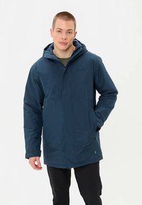 Veste imperméable marine avec capuche, dotée d'une fermeture éclair à l'avant, de poches latérales et d'une finition texturée. Le modèle porte une chemise verte en dessous.