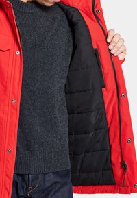 Röd ytterjacka med ståkrage, tryckknappar och ett vadderat svart foder. Den inre texturen är slät med synliga sömmar. Mörk tröja under.