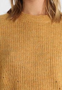 Musztardowy żółty sweter z dzianiny o prążkowanej fakturze, zaokrąglonym dekolcie i miękkim, puszystym wykończeniu. Bez widocznych ozdób ani wzorów.