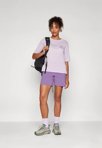 Ανοιχτό μοβ μπλουζάκι με κοντά μανίκια και μικρή γραφική παράσταση, συνδυασμένο με ανοιχτό μοβ σορτσάκι. Μαύρο σακίδιο, γκρι αθλητικά παπούτσια και λευκές κάλτσες.